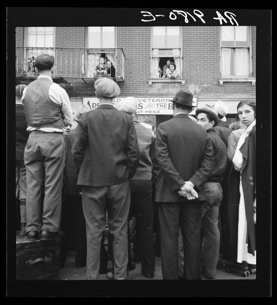 The image is a black and white photograph capturing a moment of social or political gathering. It shows a group of people, mostly men in suits and hats, standing closely together as they face the camera from behind. They appear to be waiting for something at what seems like an outdoor post office, indicated by a sign that reads "POST OFFICE HERE." Some individuals are looking up towards two women on a balcony above them who are peering down.

On the left side of the image stands a man in casual attire and jeans with his back turned; he is positioned higher than the crowd below. The focus is mainly on these people, but there's another group to the right that includes individuals wearing dresses, suggesting gender diversity within this gathering.

The photograph exudes an atmosphere reminiscent of mid-20th-century America, reflecting a historical context where public gatherings often addressed significant societal issues or civic duties like visiting post offices. The image carries weight in its depiction and conveys moments from urban life during the early years after World War II, as suggested by one caption identifying it with veterans' services at that time.

The specific location is noted to be Lower East Side, New York City; this neighborhood was known for being a hub of Jewish immigration and immigrant communities. Such an image might also prompt thoughts on collecti [...]