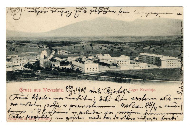 The image is a black and white postcard featuring an aerial view of what appears to be prison camp facilities. In the foreground, several large buildings with rectangular shapes are clustered together, suggestive of barracks or administrative structures within a larger complex. Among these buildings, there's one emitting smoke from its chimney, indicating some sort of activity occurring inside.

The middle ground shows more open land leading up to rolling hills in the distance under an overcast sky. The landscape is devoid of vegetation and has patches that could be fields or undeveloped areas.

On top of the postcard, handwritten text appears at various points across the image; however, it's partly faded and not entirely legible due to its vintage condition. Notably, a message begins with "Gruss aus Nevesinje," which translates from German as "Greetings from Nevesinje." Below this greeting are more cursive lines of writing that seem personal in nature.

The bottom right corner has additional text indicating the date 2/1-908 and mentions names associated with a Lager (prison camp), along with some non-Latin characters. The handwriting is quite elaborate, suggesting it may have been written by someone familiar or connected to those referenced on the postcard.

In summary, this image captures an historical snapshot of Nevesinje prison camps as viewed from above during World [...]