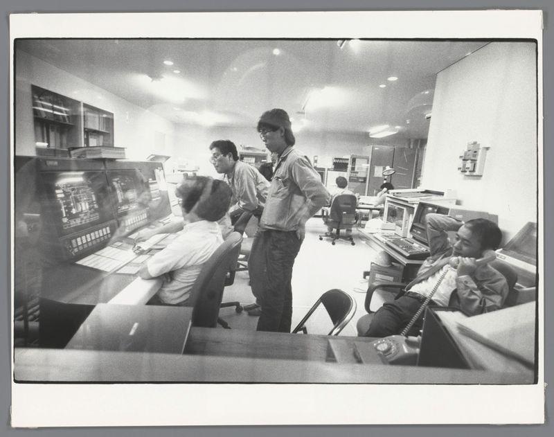 The image is a black and white photograph showing several individuals in an office setting. The central figure, likely male due to the hairstyle visible from behind, stands with his back slightly turned towards us, looking over some sort of control panel or computer monitor filled with various dials and displays. He wears glasses, has shoulder-length hair that's swept back, a light-colored shirt, dark trousers, and is holding what appears to be an adjustment knob in one hand.

Behind him, another individual leans forward attentively at the same console, wearing glasses as well and dressed similarly, suggesting they may work together or hold similar positions. To his right, a seated person with their face partially obscured by movement sits in front of another control panel, focused on something out of frame.

To the far left, an over-the-shoulder view reveals part of someone's profile: glasses-clad head and shoulders peeking from behind what could be papers or documents. On the rightmost side, a seated individual holds up their hand near the face in a gesture that might indicate thoughtfulness or fatigue, wearing dark-colored attire.

The office environment is lit by overhead fluorescent lights reflecting off surfaces with various computer terminals and monitors scattered throughout. The setting suggests an early to mid-1980s timeframe based on technology present, such as [...]