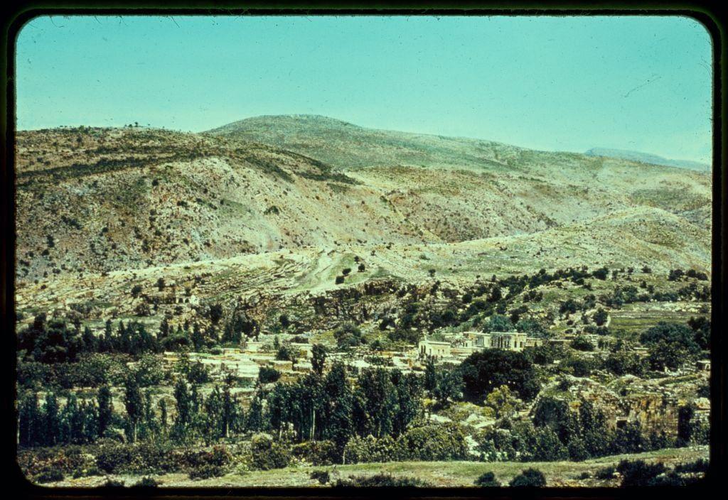 The image shows a picturesque landscape with rolling hills and terraced fields, indicative of an agricultural region. In the foreground, there is dense greenery including trees, shrubs, and cultivated plants suggesting fertile land possibly used for farming or grazing animals.

In the middle ground, clusters of buildings can be observed nestled among the vegetation; these structures seem to have a traditional architectural style with flat roofs and white walls which may suggest they are residential homes. Some larger stone constructions hint at historical ruins or ancient monuments that blend into their surroundings subtly.

The background reveals an expansive view of hilly terrain extending towards distant mountains, giving depth to the landscape while also providing context for its geographical setting within a mountainous region.

This image is framed with what appears to be vintage film grain and slight color discoloration which indicates it could have been captured on 35mm or similar photographic medium. The vignette effect at the edges suggests that this photo was taken from inside another frame, possibly during travel photography where scenes were viewed through a window or glass barrier.

The overall atmosphere of the image evokes tranquility and simplicity in rural life within a historically rich environment.