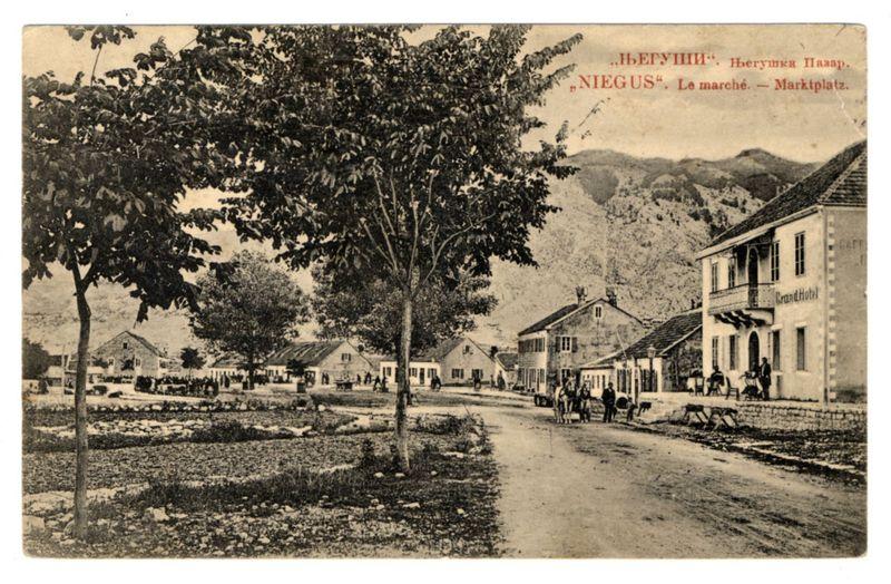 The image depicts a vintage postcard featuring an old European town. In the foreground, there's a dirt road leading through what appears to be a marketplace or main square labeled "Le marché" in French and "Marktplatz" in German. Trees line one side of the street, with houses on both sides displaying traditional architecture typical for historical periods.

In the middle ground, several individuals can be seen engaging in various activities: some appear to walk along the road while others gather near buildings that could serve as shops or residences. A group is visible by a building labeled "Grand Hotel," suggesting it's an establishment offering accommodation and possibly dining services.

The background showcases hilly terrain with more trees, creating depth for the scene. The overall atmosphere of the image suggests daily life in this small town during its heyday before modernization swept through rural areas.

On top of the postcard is text indicating a place name: "NIEGUS," which could be part of an old or misspelled reference to Niğbolu, Turkey. It's also marked with additional details suggesting locations such as Héryukh and other Russian words possibly referring to nearby towns.

The image carries historical significance, providing insight into a moment frozen in time within this quaint European locale.