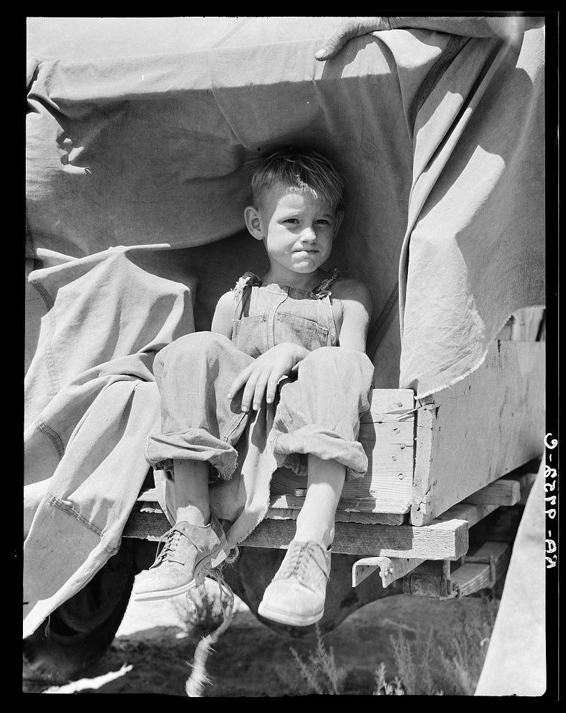 The image is a black and white photograph depicting a young boy sitting on what appears to be the back of an agricultural vehicle, possibly part of farm equipment. He has short hair, wears suspenders over tattered trousers, and his feet are clad in worn shoes. His posture suggests weariness or fatigue as he rests against a large piece of fabric draped overhead, which could indicate makeshift sheltering from harsh sunlight.

The photograph exudes an aura of poverty and hardship, likely captured during the Great Depression era when such images were widespread to document social issues like unemployment and displacement among impoverished families in rural America. The image is credited with metadata indicating it's part of a collection highlighting "a child of an impoverished family," suggesting this photo was taken by Dorothea Lange for her famous work documenting socio-economic conditions.

Additional details include the watermark text at the bottom right corner, which reads “DOROTHEA LANGE - GARDNERS,” possibly referring to Gardners Photo Service in New Mexico. The black border of the image is also part of its presentation, a common feature when prints are mounted or framed for exhibition purposes.

Overall, this photograph serves as a poignant historical record capturing the resilience and struggle faced by children from impoverished backgrounds during challenging times.
