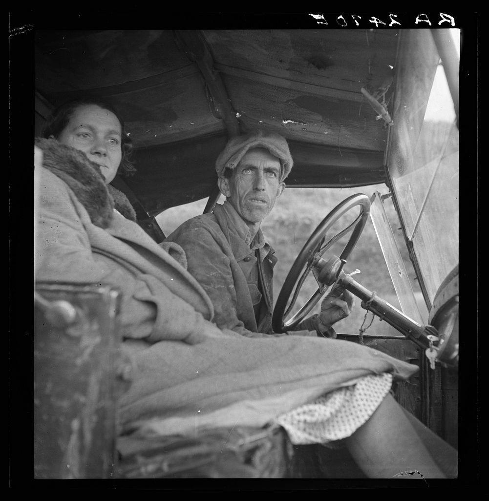 The image is a black and white photograph capturing two individuals inside an old-fashioned, open-top vehicle. The person in the driver's seat appears to be an older male with stubble on his face, wearing glasses and what seems like work attire including gloves. He is gripping a large steering wheel indicative of older automobile models. To his right, slightly reclined, sits another individual who could possibly be female based on visible clothing—a shawl or scarf—wearing darker colors which contrast against the lighter interior of the vehicle.

The quality of their clothes and demeanor suggests they may not have much wealth, potentially reflecting an era where such conditions were common. The vehicle is rudimentary with a wooden steering wheel and lacks modern safety features like a windshield or doors that close. It looks functional but worn out, suggesting it might be in use for traveling over significant distances.

The image gives off the aura of hardship and possibly migration given their attire and demeanor—traits often associated with migrant laborers during certain periods. There is visible text on the upper right corner which seems to indicate a caption or title related to this photograph but its full context isn't clear from the provided information, although it hints at something about California.

Additional details such as lighting conditions suggest that th [...]