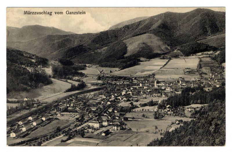 This black and white photograph depicts an expansive aerial view of a small town nestled within mountainous terrain. The landscape includes cultivated fields, patches of forestation, buildings in varying architectural styles that suggest different periods or uses, and roads meandering through the area.

The top portion of the image showcases rugged mountains with dense woodland cover, indicating hilly to mountainous geography. Below this natural backdrop is a more densely built-up section where clusters of structures are evident, suggesting residential areas interspersed with some larger buildings that could be public or institutional facilities.

A winding river cuts through one side of the town, enhancing its picturesque quality and possibly serving as an important water source for both domestic use and agriculture. The urban layout appears organized yet quaint, lacking modern infrastructure indicative of earlier times.

The image has a historical feel due to its monochromatic tone and style, reminiscent of early 20th-century photography when such aerial views were less common but gained prominence with advancements in photographic technology.

There is text at the top left corner reading "Mürzzuschlag vom Ganzstein," which translates from German as "The Mürzschlag Flood From Ganzer." This suggests that the photograph may have been taken shortly after a significant floo [...]