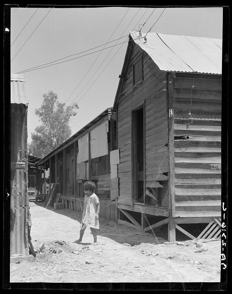 The image is a black and white photograph depicting an outdoor setting with rustic buildings. In the foreground, there's a young child wearing what appears to be a dress or robe walking on dirt ground next to one of these structures. The architecture suggests rural simplicity; wooden planks are visible in the construction, as well as makeshift supports for walls. Electrical wires run above and along sides of the building, indicating some level of infrastructure within this setting.

The environment seems arid with no signs of vegetation around except a few trees or shrubs in the background, which could suggest an agricultural area given their sparse distribution. The photograph is marked "Dorothea Lange," implying that Dorothea Lange took it and perhaps associated with its subject matter; she was known for her photography documenting life during different periods including the Great Depression.

Additional context indicates this image portrays a scene from Brawley, Imperial Valley, California concerning Mexican field laborers. The setting appears to be part of a series or collection captured by Dorothea Lange related to social conditions and lives in that specific locale at a particular time period.