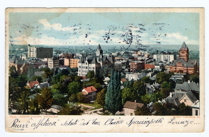 The image is a vintage postcard depicting an aerial view of the cityscape of Grand Rapids, Michigan. The scene shows various buildings with different architectural styles and colors spread across a hilly urban landscape. Prominent structures include what appears to be churches or historical edifices in red brick and white stone, alongside more modern multi-story residential and commercial buildings.

Trees are interspersed between the built environment, suggesting parks or green spaces within the city. The sky is partly cloudy with some dark spots that could indicate birds or atmospheric disturbances. A handwritten inscription at the bottom of the postcard reads "Piùvi affiliai" which translates to "More connected," followed by other text in Italian and a date stamp indicating it's from 1925.

The postcard has signs of aging, with creases, spots, and discoloration that suggest its historical significance. The bottom right corner features the Detroit Publishing Co.'s name, identifying them as the publisher or distributor for this image.