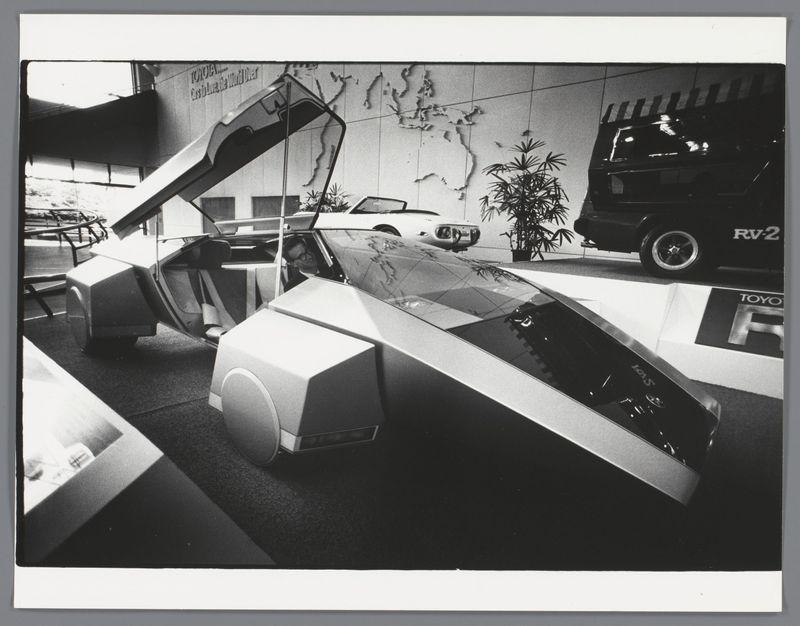 The image is a black and white photograph depicting an innovative, futuristic-looking vehicle on display in what appears to be an auto showroom. The vehicle has unconventional design features such as elongated windows with reflective surfaces that create distorted reflections of the surroundings. It also includes unique elements like a sloped ramp leading into the interior from one side, suggesting it may have no doors or conventional entry points.

In the background, other vehicles are visible, including a classic car and an RV-2 branded vehicle labeled 'Toyota.' A map is seen on the wall behind these exhibits, hinting at the global reach of Toyota's presence. The setting seems to be indoors with artificial lighting casting shadows that add depth to the scene. 

The photograph captures a moment where design innovation meets consumer automotive display, reflecting on how vehicles were envisioned and marketed in Japan during the late 1980s or early '90s era when Ed van der Elsken was active as an artist.

Additional context about this image can be found through Auto in een Toyota showroom in Japan by Ed van der Elsken between 1985-1990, with a specific reference to the photograph numbered 6484.