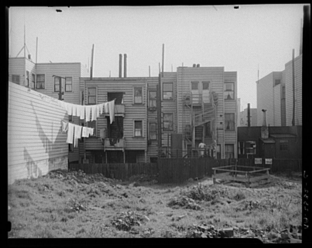 The image depicts an urban scene from the early 20th century. It shows a row of wooden buildings with flat roofs and various features such as balconies, air conditioners, and utility lines visible on their exteriors. The buildings appear to be residential housing or multi-family dwellings, likely in a working-class neighborhood.

A clothesline is stretched across the street with laundry hanging out to dry, suggesting daily domestic life activities taking place within this community space. In front of the building closest to the foreground, there's an open area covered with grass and weeds, indicating possible neglect or lack of maintenance. A wooden fence separates part of this unkempt ground from a cleared lot where no visible structures stand.

The black-and-white photograph conveys a sense of historical context, possibly documenting living conditions in San Francisco during that era as noted by the source provided (https://images.loener.nl/DorotheaLange/full/6495/6495b85fba25398d90c506b1.jpg). It reflects a candid snapshot of urban life in America's past, with an emphasis on the social fabric and architectural style prevalent at that time.
