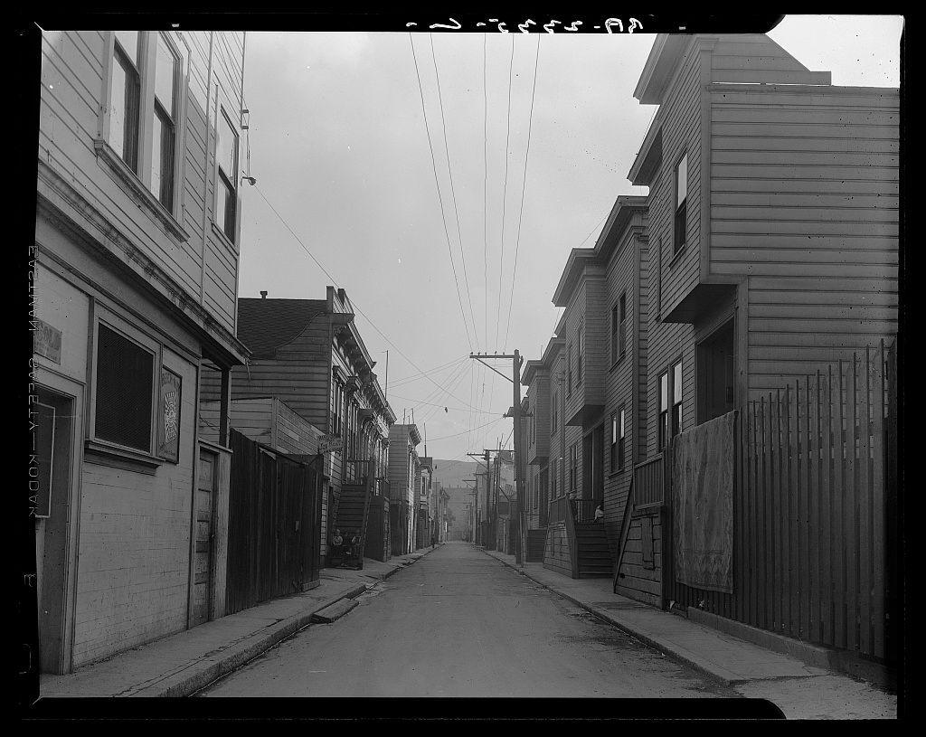 The image is a black and white photograph depicting an urban alleyway, likely from the early to mid-20th century. The scene shows two rows of wooden housing structures with pitched roofs on either side of the narrow street. Electrical power lines run parallel overhead between the buildings.

On one side, there's visible wear and damage; some signs are faded or peeling, indicating age or neglect. One building displays a banner that reads "FIRE DEPT." hinting at historical use as emergency housing for firefighters during fire season. The alley is devoid of people but has scattered objects like chairs on the sides which suggest habitation.

The street itself appears to be paved and in decent condition with no visible cars, suggesting it may not have been a primary thoroughfare or could represent a period when such streets were more prevalent before auto dominance. In the background, an open space is seen that looks into another hilly urban landscape typical of San Francisco's terrain.

This photo seems like part of Dorothea Lange’s work in documenting living conditions during and after the 1906 earthquake, capturing real-life scenes to advocate for housing reforms or social justice. The caption suggests a connection to Mission District, likely highlighting how residential spaces were utilized post-disaster under challenging circumstances.