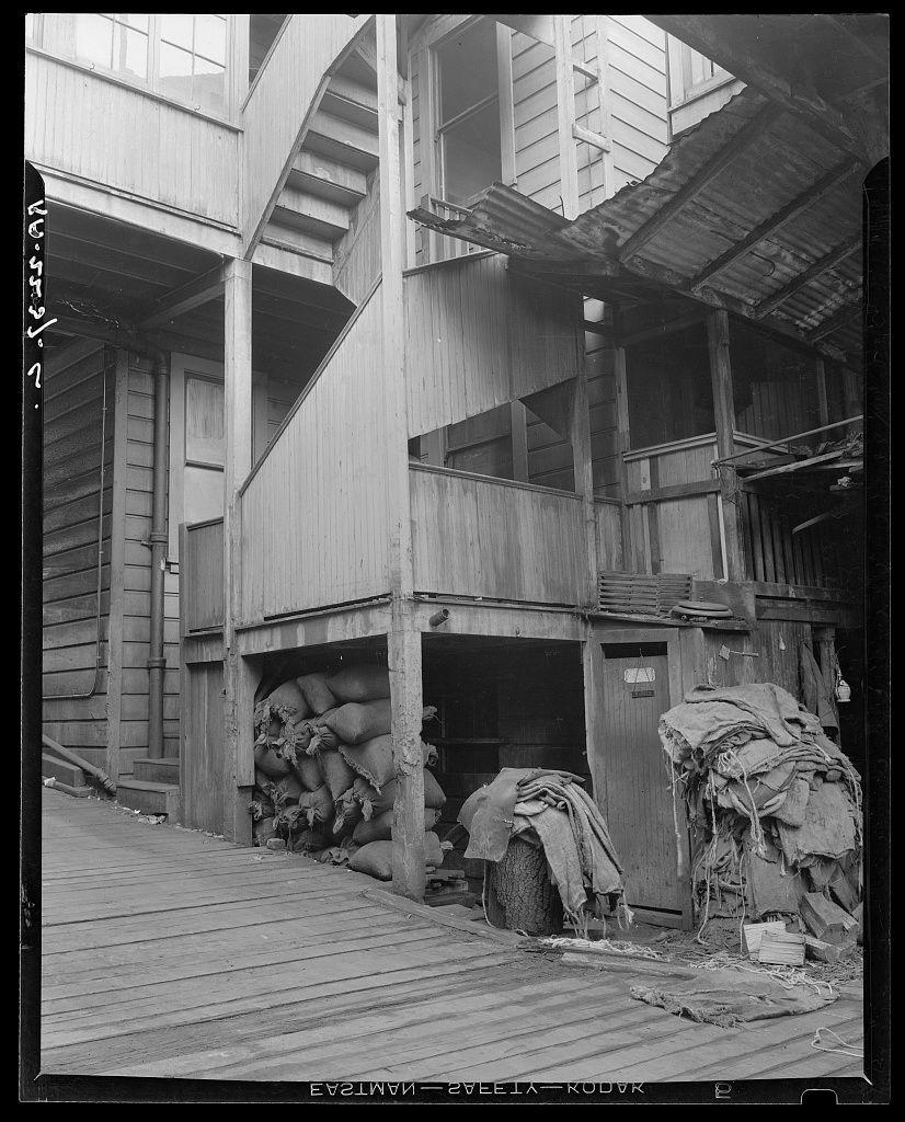 The image depicts a monochromatic scene of an industrial setting with wooden structures and various textiles or sacks arranged in different parts. The architecture suggests multiple levels, possibly for storage or processing purposes. There are visible elements like pipes along the building's facade and what appears to be fabric or clothing items hanging on racks outside one section. Notable features include a clear view down into an open space beneath some of the wooden structures where more textiles are stored in burlap sacks.

The environment has an aged appearance, with signs of wear such as peeling paint and exposed wood beams. The flooring is made up of wooden planks that show significant use over time. There's no visible human presence or modern technology indicating a historical context for the photograph. This scene likely represents part of North Beach District in San Francisco during a period where Italian American immigrants were prominent, hinting at cultural influences from Italy.

The image carries an air of nostalgia and abandonment, possibly illustrating a once-bustling area now left to history's remnants. The absence of vibrant colors accentuates the texture contrasts within the scene, emphasizing worn materials against raw wood structures.