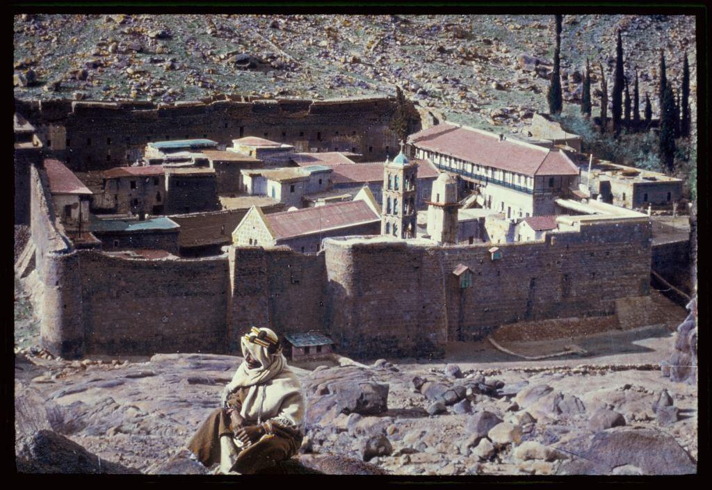 The image depicts a historical and arid landscape, featuring an ancient walled settlement with multiple buildings that include what appears to be a church or monastery. The architecture suggests Middle Eastern influence, characterized by stone walls and terracotta rooftops.

In the foreground, there is a figure dressed in traditional attire resembling a desert dweller, sitting on rocky ground holding a jug-like container, possibly for water storage. This person seems contemplative or observant of their surroundings.
The background reveals a mountainous terrain with sparse vegetation primarily consisting of cacti and shrubs. The overall color palette consists of earthy tones such as browns, reds, and greens, which evoke the dry desert environment.

This image captures a moment in time at the Monastery of St. Katherine's close view between 1950 to 1977, according to additional information found on Sinai. The photograph is credited to Matson Photo Service from their collection, as noted by a reference number and URL provided for more details.