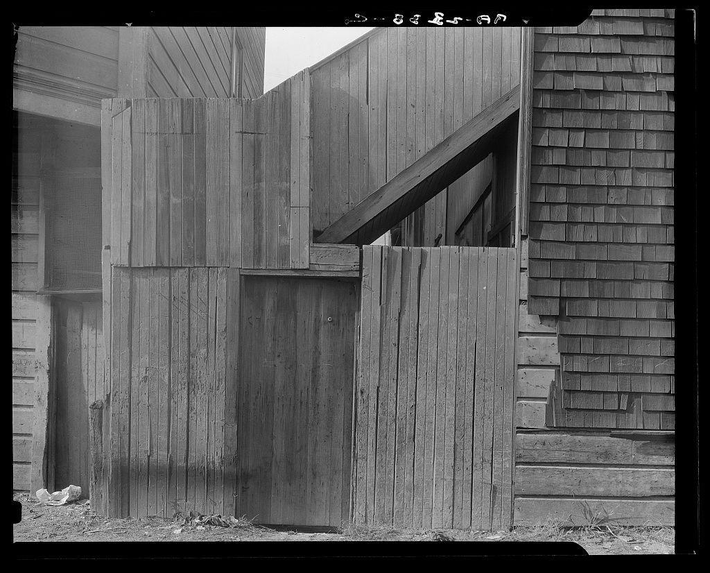 The photograph displays a weathered wooden structure with irregularly shaped and uneven surfaces. It appears to be part of an older building, showcasing elements like vertical planks in varying states of wear and a slanted roof that suggests age or disrepair. The texture is rough and the wood's natural grain pattern is visible through black-and-white tones captured by Dorothea Lange during her work on Mission District firetrap projects in San Francisco, California.

The setting appears unkempt with debris scattered near the base of the structure. No people are present in this image which focuses entirely on the architectural elements and their environmental context. The absence of color emphasizes textures and structural details while highlighting historical or socioeconomic conditions that might be reflected through Lange's lens during her documentation period around 1938-1942, as indicated by the date stamp visible at the top right corner.