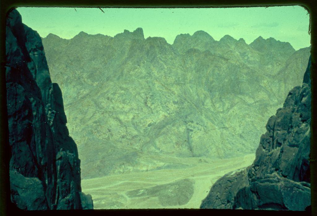 The image depicts a stunning view of an arid mountain landscape, possibly captured from within or through a window. The rocky terrain stretches far into the distance under clear skies, with rugged peaks creating a dramatic silhouette against what appears to be a bright greenish-blue sky. A winding road can be seen in the foreground, providing a sense of scale and leading toward the mountain range that dominates the view.

In terms of context, this photo seems to have been taken between 1950 and 1977, indicating it captures historical significance within the Sinai region. The image is part of an archive from Matson Photo Service, known for documenting various landscapes across different continents during their travels in Egypt's Sinai Peninsula.

This specific photograph likely has ties to biblical history as well; Jethro's path mentioned could refer to a route taken by Jethro (Moses' father-in-law), which led through the mountains of Midian. Aaron is also associated with this region, known for his role within Moses and Israelite religious traditions.

Overall, the photo offers an evocative glimpse into the stark beauty and historical weight of the Sinai landscape, blending geological features with layers of cultural significance.