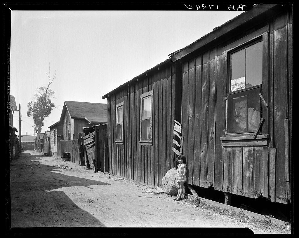 The image depicts a black and white photograph of an old railway car on tracks. The surrounding area appears to be in disrepair, with dilapidated wooden buildings lining the train track. A small child is seen sitting next to one of these structures.

This scene reflects historical living conditions during the early 20th century when communities were formed along railroad lines for economic opportunities related to rail transport and industry. The image serves as a powerful visual representation of the socio-economic challenges faced by families who lived in close proximity to train tracks, highlighting issues such as inadequate housing, poverty, and displacement.

The photograph has been identified with Mexican quarter of Los Angeles, located one-quarter mile from City Hall. It is mentioned that this area was condemned due to its deteriorated condition and slated for demolition soon after the construction of a new Union Railroad station would take place in the vicinity. The average rent during that time period averaged around eight dollars.

The image captures both the historical context as well as serves as an illustration of social inequality, providing valuable insights into urban living conditions from over a century ago.