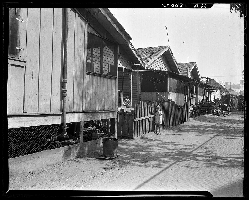 Black and white photograph depicting a street with wooden houses. The scene appears to be from an earlier time period, judging by the style of housing and clothing worn by two visible individuals - one on a raised platform beside a house's side entrance, likely performing laundry or cleaning work; another child standing in front of a fence closer to the camera holding what seems like a toy. There are various objects such as barrels, metal buckets, pipes extending from houses, an elevated wooden structure with steps leading up to it possibly for children play, and a clothesline strung between poles bearing hanging garments. The street is unpaved or has dirt surfaces, without any visible modern vehicles, suggesting this photo could be circa early 20th century based on the architectural style of housing and lack of paved roads typical in urban settings today.