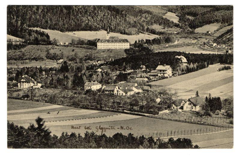 The image is a black and white photograph depicting an expansive rural landscape with various buildings, fields, trees, and hills. In the foreground, there are large agricultural plots arranged in neat rows or patches of different shades, suggesting crop cultivation. To the left side, we see what appears to be several small houses clustered together on elevated ground, surrounded by bare trees indicating a colder season.

In the middle distance, various residential buildings with pitched roofs and chimneys dot the landscape against rolling hills covered in sparse vegetation and leafless deciduous trees indicative of winter or early spring. The most prominent structure is a large building resembling an estate or manor house located on higher ground amidst these houses, signifying its importance.

On the right side of the image, more residential buildings are visible along with what seems to be vineyards in rows, suggesting viticulture practices. In the far background, densely forested hills rise against the horizon under a clear sky. The photograph is annotated at the bottom left corner with text that reads "Hart bei Gloggnitz - N.Oe," which translates from German as "Gloggnitz near Vienna."

The overall composition of this image conveys an overview or panoramic view of rural life and agricultural practices in what appears to be a European setting, potentially Austria give [...]
