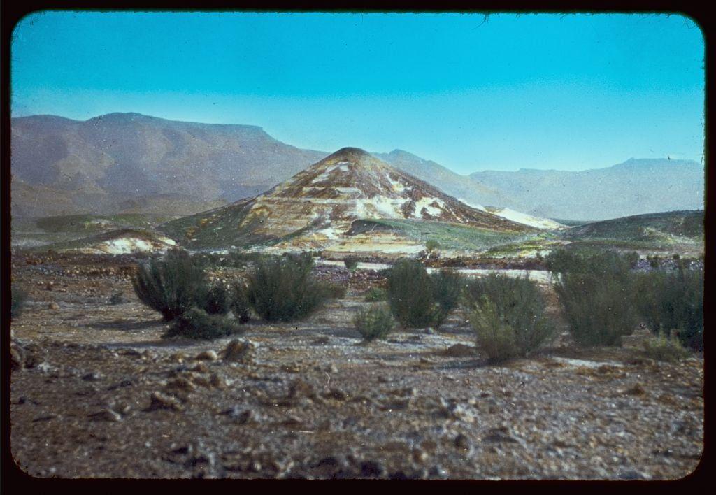 The image depicts a barren, desert landscape with sparse vegetation. In the foreground, there are small shrubs and rocky terrain indicative of arid conditions. The middle ground is dominated by a prominent hill or mountain covered in patches that appear to be mineral deposits or lagoons, creating contrasting colors such as yellowish-brown and white against the greenery at its base. This geological feature stands out prominently against the backdrop.

The background features distant mountains with varying shades of brown and gray, suggesting their rocky nature and distance from the viewpoint. The sky is a clear blue without visible clouds, implying fair weather conditions typical for arid regions like those in Sinai during that time period (1950-1977). Overall, this scene captures an environment characteristic of mountainous desert landscapes found in Egypt's Sinai Peninsula.

Additional information about this image can be accessed through the source link provided.