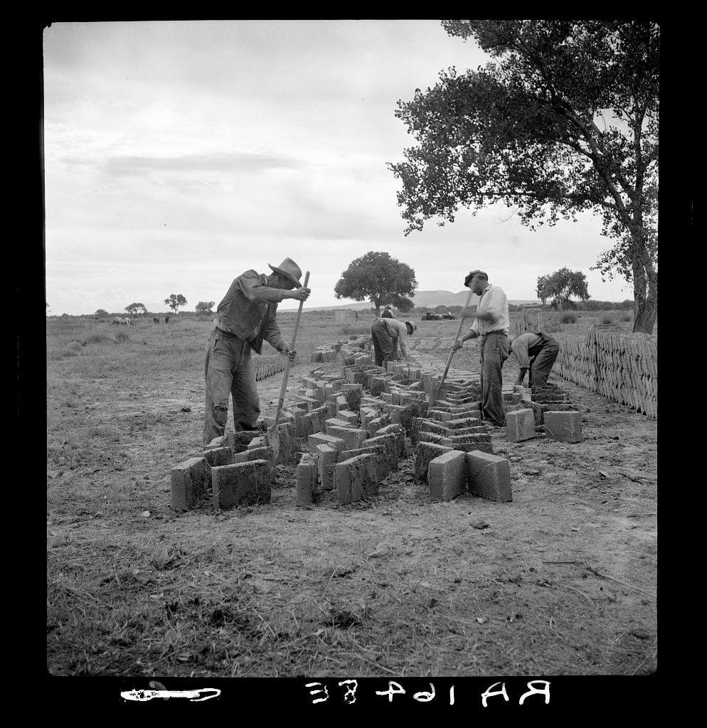 A black and white photograph shows a group of people in an outdoor setting, working together to shape and stack rectangular concrete blocks. They are using tools such as hoes or shovels to handle the material and construct what appears to be rows of stacked bricks. The environment is rural with trees visible in the background and open fields extending into the distance under a cloudy sky. There's also some fencing along one side, suggesting that this work may take place on farmland or ranch property.