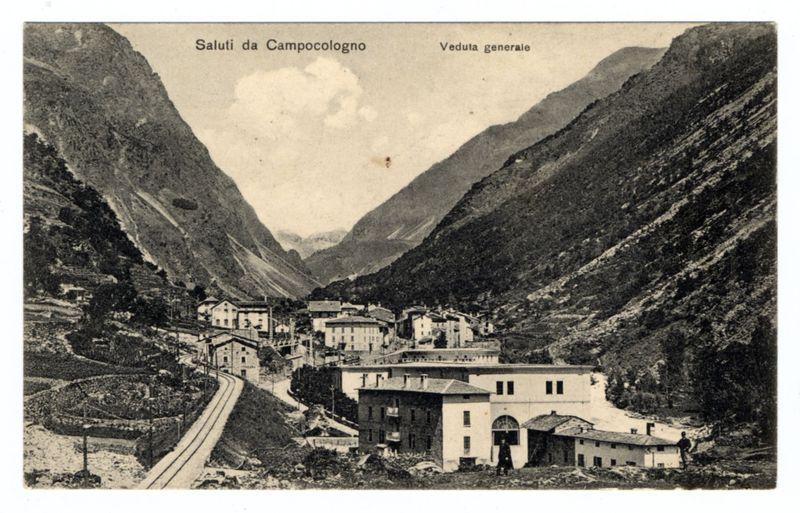 A vintage black and white photograph depicting a picturesque mountainous landscape. The view showcases the Saluti da Campocologno, as indicated by text at the top of the image. In the center is an old village with multiple buildings featuring traditional European architecture, including houses and what appears to be communal or public structures like schools or churches.

The scene is dominated by steep hills on either side filled with sparse vegetation and rugged terrain that suggests a high-altitude environment. A railway track runs parallel along the valley floor from left to right, indicating transportation infrastructure in this remote area. The village sits at an intersection between two tracks which may be part of a train line.

Above the photograph is text "Saluti da Campocologno" and below it reads "Veduta generale," both likely referring to greeting or general view related to Camptocolo, possibly Italy given its linguistic similarity. The overall atmosphere evokes an early 20th-century setting with a historical feel due to the sepia tone of the image.

The bottom part of the photograph shows some debris and rubble, suggesting possible damage or disrepair within this village context, which might imply that this photo was taken during reconstruction after natural disasters like earthquakes.