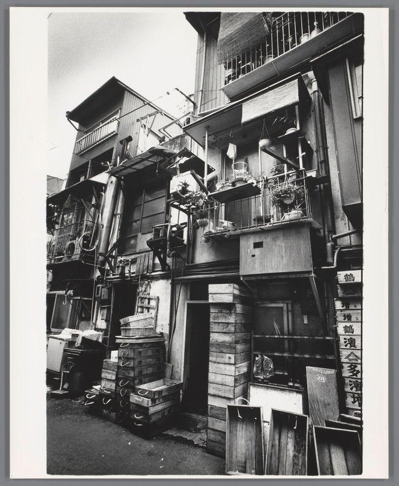 This black and white photograph captures the intricate details of a multi-story building's exterior, indicative of urban living in Japan during the late 1950s to early '60s. The image is densely packed with architectural features: wooden balconies adorned with potted plants, exposed piping on external walls, various signs bearing Japanese characters, and numerous stacked wooden crates suggesting a bustling neighborhood possibly involving commerce or industry. Balconies are furnished sparsely yet reflect an attempt at personalization within limited spaces. A sense of clutter is present due to the disarray in which items such as electrical equipment and laundry hang from lines across balconies.

The composition draws attention upward, emphasizing the height and density typical of city environments during that era. The absence of people gives a still life quality to the scene, inviting viewers to ponder on daily lives amidst urban living. The photograph is rich with texture—wooden planks, concrete walls, metal pipes—all contributing to a tactile narrative of the environment.

The use of monochrome adds an element of timelessness and focus on form over color. It's reflective not only of photographic technology constraints at that time but also serves as a stylistic choice by Ed van der Elsken for his work in Japan. The photograph is part of 'Achterzijde' (Backside), series, h [...]