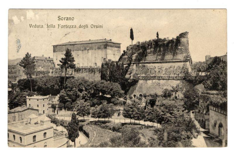 The image is a sepia-toned vintage photograph depicting an aerial view of Sorano, highlighting the "Veduta della Fortezza degli Orsini," or View of the Fortress of the Orsini family. The fortress appears prominently on elevated ground with its walls and defensive structures visible against a backdrop that includes another large building to its left.

In front of these imposing architectural features lies an area abundant in greenery, consisting mainly of trees and some structured garden areas where people can be seen walking or standing. To the lower-left foreground is a smaller, simpler structure with what seems like a rooftop terrace adorned by one lone tree. The overall scene suggests historical significance coupled with elements indicative of daily life.

The photograph carries an aged appearance typical of early 20th-century prints and includes visible text in Italian indicating that it documents Sorano and specifically the Fortress Orsini. It is likely part of a collection or publication, given its styled presentation on what appears to be paper stock commonly used for postcards or similar printed materials at the time.

The image captures an era gone by but also provides insight into how historical sites were documented in print media during that period. The photograph serves as both a piece of history and a visual record of cultural heritage, reflecting Sorano's p [...]