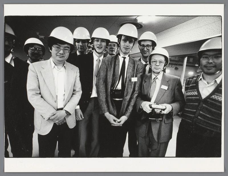 The image is a black and white photograph featuring a group of men, likely taken during the 1980s or early 1990s given its monochromatic style. The individuals are standing close together in what appears to be an indoor setting with reflective ceilings that suggest it might be above a subway station or within a similar infrastructure.

In the foreground is a man wearing glasses, dressed in business casual attire consisting of a blazer and trousers, paired with a light-colored shirt underneath his jacket. His hands are clasped in front, suggesting he may not want to draw attention directly towards himself but rather as part of the group.

Surrounding him on all sides are men who wear white safety helmets, indicating that they might be involved in construction or some form of industrial work. They display a mix of casual and formal attire, with most wearing jackets and one individual notably holding what appears to be a walkie-talkie or communication device under his arm.

The expressions range from neutral looks towards the camera to more animated smiles among several men, suggesting a positive atmosphere despite their professional setting.

Additional information about this image can be found in Portret van Groningse studenten en mannen van een organisatie in Japan by Ed van der Elsken. The reference suggests that these individuals may have connections with students from  [...]