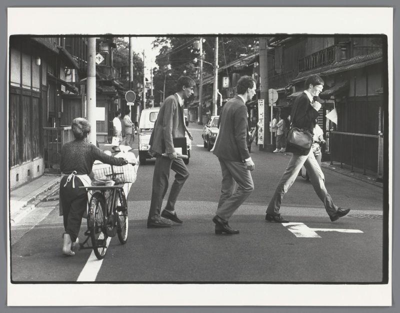The image captures a moment on an urban street, likely in Japan given the context provided. It is rendered in black and white, which adds to its timeless quality. In this still life of daily life, we see several figures crossing at what appears to be a pedestrian crosswalk marked by distinct white stripes.

On the left side of the frame, there's an elderly woman with her hair pulled back into a bun, pushing a bicycle laden with goods across the street. Her attire is casual yet practical for running errands; she wears dark trousers and carries a small bag over one shoulder while wearing what appears to be rubber boots or shoes.

In contrast, on the right side of the frame are three men who seem engrossed in their own activities as they traverse the crosswalk. The man leading seems intent on walking with purpose, possibly heading towards work given his formal attire—a blazer and trousers paired with a light-colored shirt—and carrying what looks to be a briefcase.

The second figure follows closely behind him; he is also dressed formally but without accessories like glasses or earphones that could suggest he's in transit for professional reasons. The third man, slightly ahead of the other two, has his head lowered and appears lost in thought or perhaps listening to something through headphones—details hinting at a more relaxed pace.

The street itself hints at an urban setti [...]