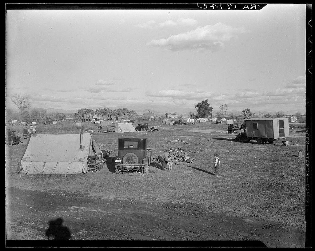 This black-and-white photograph depicts an outdoor setting with a makeshift settlement consisting of tents, trailers, and other temporary structures. The scene suggests the presence of a community living in difficult conditions, possibly during or following a period of displacement or economic hardship.

In the foreground on the left side is a large tent-like structure made from various materials including canvas. Adjacent to it stands an older model truck with its back opened up as if used for storage or transport purposes. Scattered around this central area are several people, some standing while others appear engaged in tasks such as carrying objects.

The middle ground reveals more of the settlement; a row of trailers and additional tents can be observed. The background shows open land leading to hills under a partly cloudy sky. There's no visible vegetation close by, which might imply that this area is not well-established for agriculture or habitation.

On-site activities seem sparse as people are few in number compared with the expanse covered by makeshift shelters and vehicles used likely for daily sustenance tasks like cooking food over an open fire seen on the right side. The overall atmosphere appears somber, reflective of a community coping under challenging circumstances without access to stable housing or resources.

The photograph’s historical context is su [...]