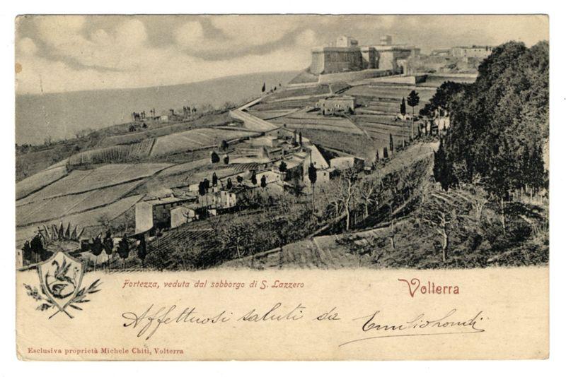 The image depicts a sepia-toned photograph of an old postcard, showcasing a panoramic view of the cityscape and landscape in Volterra. In the foreground is a large fortress with multiple towers standing prominently on elevated ground overlooking expansive cultivated fields or vineyards stretching out to meet the sea at the horizon line. The terrain appears hilly with patches of wooded areas interspersed throughout.

The architecture of the fortress suggests historical significance, possibly from medieval times. It stands as an imposing structure against a backdrop where clouds gather in the sky above the coastline. Below and around this fortification is a mix of small buildings that seem to form part of what could be residential or commercial quarters associated with the fortress complex.

Prominently displayed at the top left corner, there's text indicating "Fortezza, veduta dal sobborgo di S. Lazzero," which translates from Italian as 'Fortress, viewed from the foothills of St. Lazarus.' The bottom right features another label reading "Volterra" in stylized red lettering.

The postcard also contains handwritten cursive text and a signature that likely represents personal inscriptions or annotations made by someone who either visited Volterra or has ties to it. These details provide a sense of intimacy, as if the card might have been sent from afar with heartfelt sentime [...]