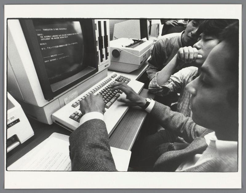 The image is a black and white photograph depicting two individuals engaged with vintage computer equipment. One person, wearing a suit, is seated at the keyboard of an early model desktop computer displaying text on its monitor, suggesting some form of data entry or programming activity. The second individual appears to be in deep contemplation, resting their head on their hand near another piece of electronic machinery that looks like either a printer or copier.

The setting seems informal and possibly indicates collaboration between the two individuals. The technology used is not modern by today's standards; it features bulky monitors with a distinct 1980s design aesthetic for personal computers, which implies this photograph was taken during that era. There are documents on the desk in front of them, further indicating an academic or professional context.

The image carries historical significance as it captures early computer use and human-computer interaction before widespread accessibility to such devices today.