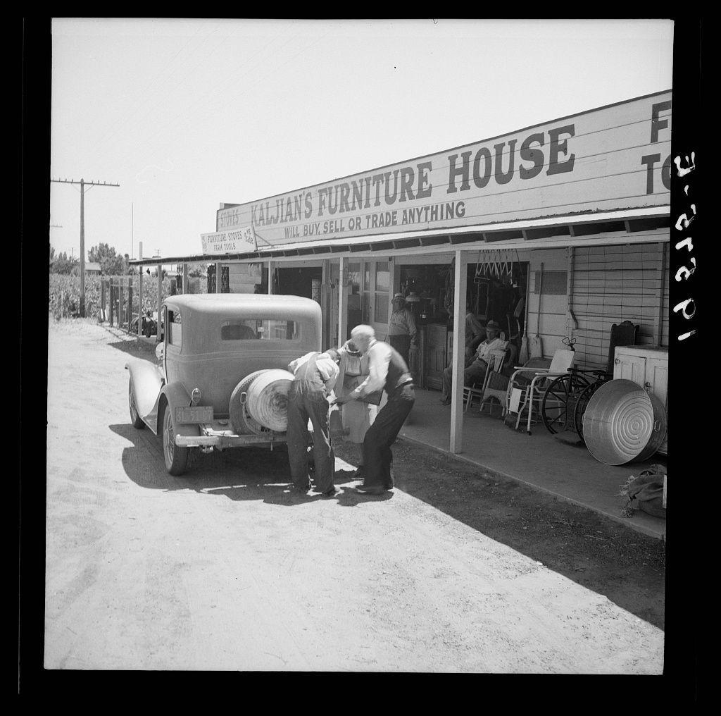 This black and white photograph captures a moment from what appears to be the early 20th century, featuring two individuals working with large rolls of material next to an old-fashioned automobile. The setting is outside Kaljian's Furniture House, as indicated by the signage on top of their storefront. One individual stands beside the car while another person assists in handling the rolls.

The environment suggests a rural or semi-urban area, potentially along U.S. Route 99 near Fresno, California, based on contextual clues from other works attributed to Dorothea Lange and her photographic series documenting life during that era. The presence of outdoor furniture items like buckets adds to the rustic ambiance of the scene.

The photograph reflects a snapshot of daily life and commerce in America's heartland at the time, with small business owners actively engaged in their trade practices on the outskirts of major cities such as Fresno. Lange was known for her extensive work documenting social issues during that period, including rural poverty and agricultural labor conditions.