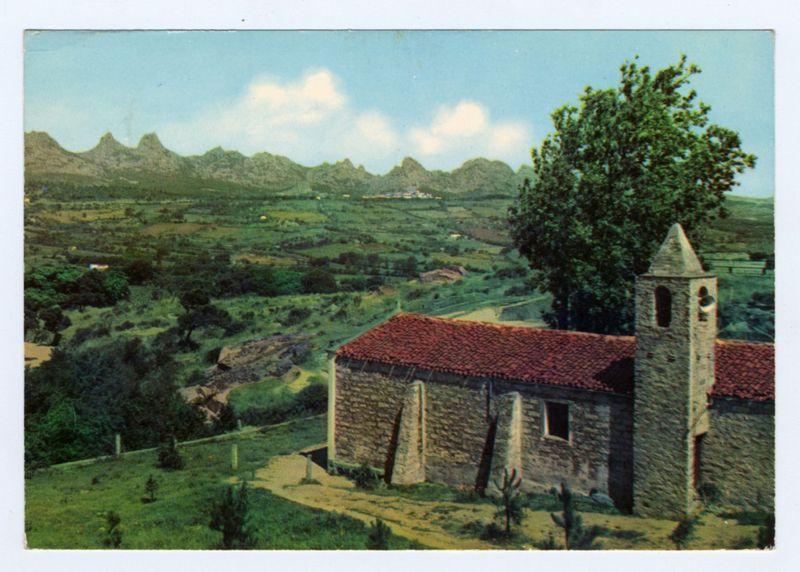This image is a vintage postcard featuring an idyllic rural landscape with a small chapel. The chapel, constructed of stone and topped with a red tiled roof, stands prominently in the foreground on the right side. It has a modest bell tower attached to one corner, showcasing arched windows typical for ecclesiastical buildings.

Behind the church, a lush countryside stretches out with fields that appear cultivated but not overly intensive farming is evident due to the patchwork of greenery and earth tones indicative of crops or pastureland. A few small structures are scattered across these farmlands, suggesting modest human habitation in this rural setting.

The midground reveals rolling hills covered sporadically by trees interspersed with more open fields, leading up towards a range of larger mountains on the horizon. These mountain peaks have distinctive silhouettes against a partly cloudy sky, hinting at either late afternoon or early morning light due to their soft shadows and warm colors.

The overall color palette is rich in earthy tones - greens, browns, reds from the chapel's roof, with hints of blue from the distant mountains. The scene exudes tranquility and pastoral beauty, characteristic of rural Italian landscapes during a time when such postcards were popular souvenirs or means for long-distance communication.