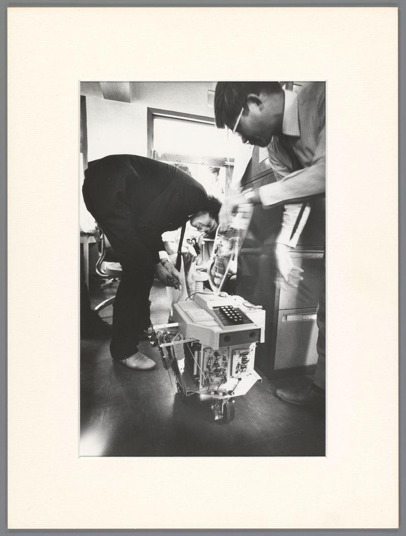 The image captures a moment within an indoor setting, likely a laboratory or workshop. A person is bent over and appears to be working on a mechanical device placed on the floor. This device resembles early robotics with visible electronic components and wires exposed at its base, suggesting it may not yet have been encased in casing for protection.

The individual is dressed casually in dark clothing, including trousers, indicative of an informal work environment or perhaps protective wear commonly used during electronics-related activities to prevent accidental damage from static electricity. The person's stance suggests focus on the task ahead with their hands actively engaged with what seems like a control box that sits atop the device.

In the background, another individual is partially visible, seated and observing attentively but not directly interacting with the mechanical creation or its operator. This second figure adds to the narrative of collaboration or observation in this technical endeavor.

The room appears utilitarian with plain walls, fluorescent lighting which casts a stark white glow over the scene, creating high contrast between light and shadow areas on the floor and subjects. The black-and-white tone enhances the timeless quality of the photograph, evoking an atmosphere that could be from any era but aligns closely to mid-20th-century aesthetics.

T [...]