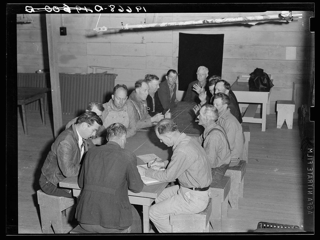 The image captures a group meeting in what appears to be an old, modest indoor setting. A dozen individuals are gathered around a large rectangular table engaged in discussion or collaboration. The room is sparsely furnished with wooden walls and flooring that give it a rustic feel.

On the left side of the frame, three men sit closely together; two lean on their elbows as they converse, suggesting an informal but earnest exchange. In contrast, another man across from them, dressed differently in what looks like overalls or a light-colored jacket, holds his hand up to emphasize his point during conversation.

The majority of participants are seated along the right side and behind the table. Their attire varies; some wear dark jackets while others sport lighter colors such as beige and white shirts with suspenders. A few individuals appear animated in their gestures, adding dynamism to what seems a serious discussion or planning session.

Notable features include the wooden benches for seating that are simple yet sturdy, reflecting practicality over aesthetics. Above them runs an exposed conduit of electrical wiring which implies basic utilities without any modern conveniences visible.

The overall atmosphere is one of community and purpose among these individuals who could be camp council members given their focused engagement in a communal setting associated with labor o [...]