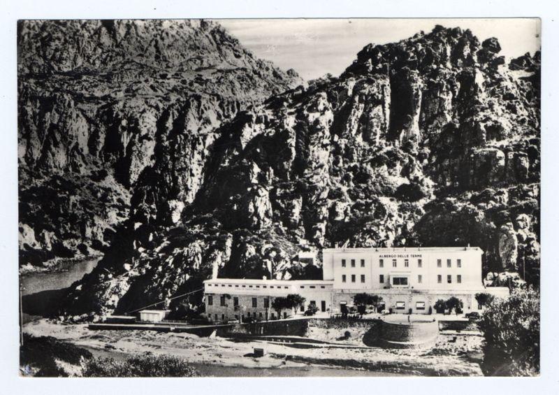 The image is a black and white photograph depicting an old, stately building named "Albergo delle Termi," nestled amidst rugged mountains. The structure appears to be composed of several connected pavilions with multiple windows, some set in arched openings typical of Italian architecture. A prominent feature is its classical facade: symmetrical arrangement, central entrance flanked by two sets of double doors and a series of evenly spaced rectangular windows.

To the left foreground lies what seems like an ancient cistern or well structure surrounded by shrubbery, hinting at historical use for water storage in this arid locale. In front of the building is expansive open space leading down to a river flowing through rocky terrain with sparse vegetation—a stark contrast against its built surroundings.

The mountains loom majestically behind and alongside the hotel, covered sporadically by trees that dot their rugged surface, adding texture but not obscuring much sunlight or view. The image conveys an aura of solitude and tranquility far from urban noise. The absence of modern vehicles suggests this might be a historical photo dating back to early 20th century or later based on architectural style and lack of contemporary elements.

No human figures are visible in the scene, which enhances its timeless quality with no distractions from the natural beauty and the grandeur of [...]