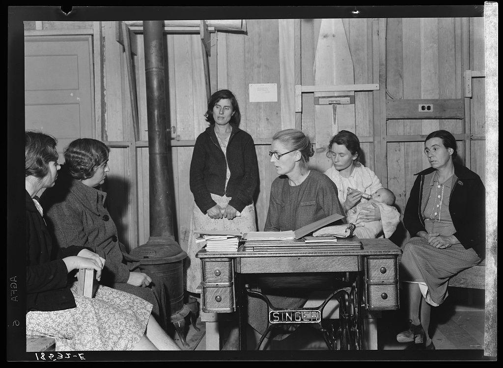 The image shows a group of six women gathered around a wooden table in what appears to be an indoor setting. The room has unfinished wood walls, and there is visible piping on one wall. One woman stands behind the others while five are seated or standing at various positions near the table.

Among those sitting down, two hold books with "SINGER" printed on them, suggesting they may involve sewing machines or related activities. A baby sits in a woman's lap to the right of the group. The women appear engaged in conversation and possibly discussing something important as indicated by their attentive postures.

The photograph is black-and-white, adding a historical context that suggests it could be from an earlier time period. Based on additional information provided (Meeting of the Mothers' Club in Arvin camp for migrant workers), this image likely captures a moment during a meeting organized to discuss practical matters affecting these women's lives as part of their community or support network within a farm labor environment.

The specific discussion topic, centered around buying and distributing kerosene oil cooperatively among themselves, highlights the economic challenges faced by migrants working on farms. This context underscores the social issues surrounding migrant workers' living conditions during that period in American history.