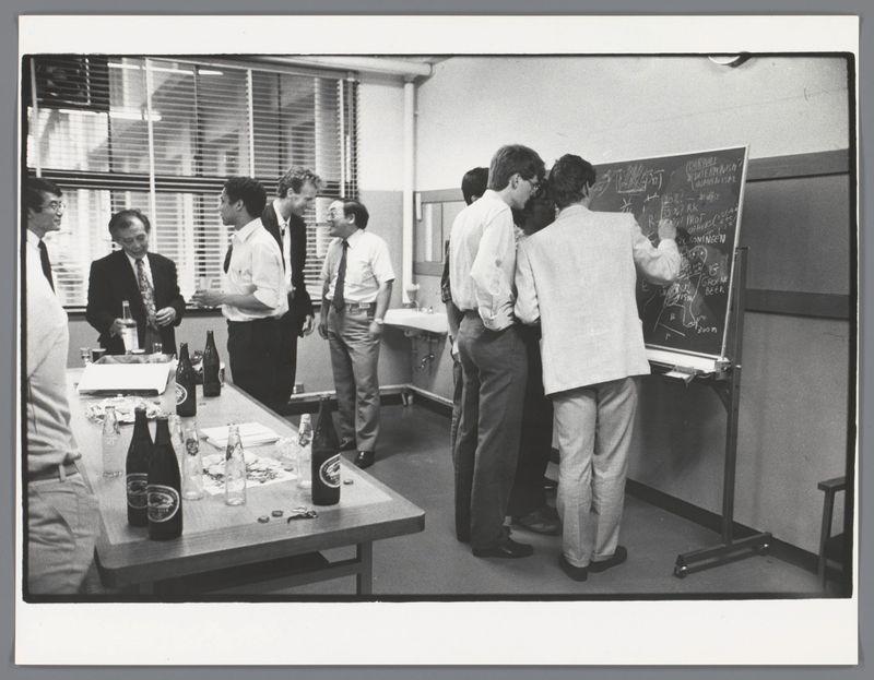 The image is a black and white photograph depicting an indoor scene, likely within an office or meeting room. A group of individuals, predominantly men in attire suggesting business casual to semi-formal dress code—such as shirts and trousers—are gathered around various tables and standing near chalkboards with writing on them, indicating some form of collaborative activity or brainstorming session.

Several people are engaging directly with the boards; one person appears to be actively writing while others observe. The presence of multiple bottles and glasses suggests that this gathering may have a social component as well, potentially involving drinks during their work discussion.

The environment is illuminated by natural light filtering through blinds on what seems like windows out of frame, creating contrasted areas in the room. There are various objects scattered across tables including papers, glassware with beverages (likely alcohol), and possibly some snacks or food items—indicating a relaxed working atmosphere.

Notably, the writing on one chalkboard appears to be written by someone who is right-handed given their stance and orientation relative to the board; however, due to the angle of the photograph this cannot be determined conclusively. The overall ambiance suggests an informal yet focused group interaction possibly related to professional or academic endea [...]