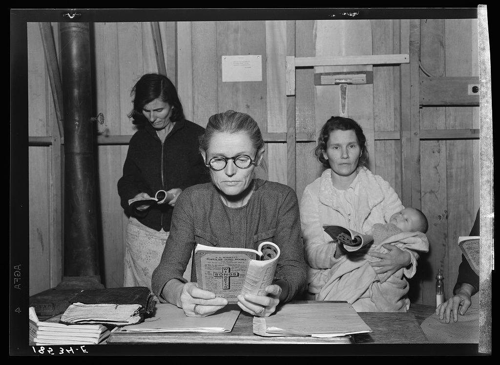 A black and white photograph shows three women seated around a table in what appears to be an indoor setting, possibly within a temporary housing structure. The woman on the left is standing with her head down as she looks at something out of frame. She wears dark clothing including a jacket over a dress or skirt. In front of this figure sits another woman who seems engrossed in reading from a booklet titled "Mother's Service Record." This seated woman, wearing glasses and dressed casually, holds the book open with her left hand while using her right to point at something within it.

On the far right is yet another woman; she is holding an infant wrapped snugly. The baby appears relaxed on its mother’s lap as they both look directly into the camera's lens. In front of them lies various papers, a bottle, and other objects suggesting daily living activities or perhaps record-keeping for their stay.

The background features wooden panels with visible signs of wear or construction elements like metal rods affixed to it. A sign is partially legible on one such rod but its content isn't clear from this angle.

This image evokes the atmosphere of a period when photographs were used not just as personal mementos, but also perhaps for documentation purposes related to social services and migration narratives during challenging times in American history.