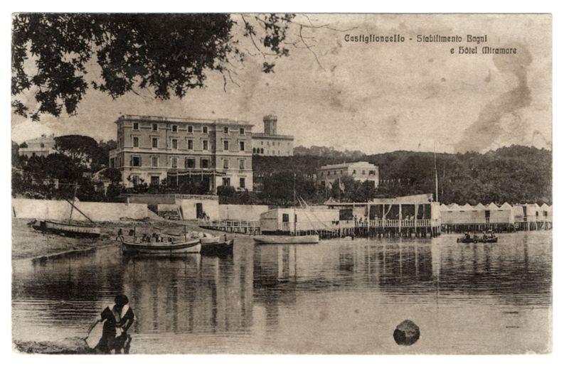 The image is a sepia-toned, black-and-white photograph depicting an early 20th-century waterfront scene. It shows calm waters with various boats anchored along the dockside in front of buildings and greenery on hills rising behind them.

On the left side stands a large building labeled "Casiglioncello," which appears to be a hotel or public edifice, possibly connected to the beach facilities mentioned due to its proximity to what seems like bathing areas. The text at the top identifies this location as Casiglioncello with references to Bagni (bathing establishments) and Hotel Miramare.

In the foreground on the left bank of water is a couple engaged in an activity by the shore, likely cleaning or collecting items from the ground. A child accompanies them. The overall atmosphere suggests leisure activities associated with seaside recreation during that era.

The image carries historical connotations as it resembles postcards and could have been used for promotional purposes to showcase the beauty and appeal of Casiglioncello's beaches and hotel offerings, thereby attracting tourism.