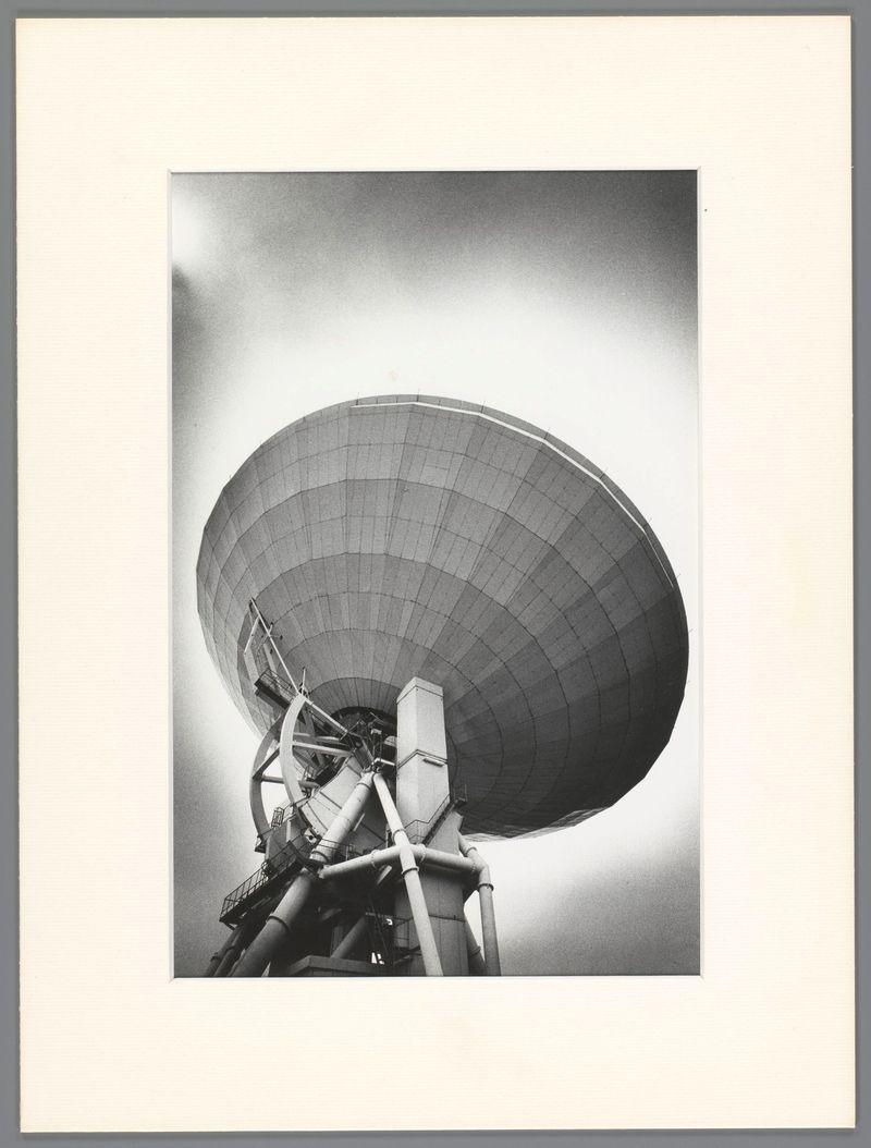This image is a black and white photograph depicting an imposing radio telescope, known as the "Schotelantenne" in Japan. The structure stands tall with its large dish-like antenna angled upwards against what appears to be a light gray sky or perhaps foggy weather conditions. It's mounted on multiple supporting structures that include various pipes and platforms, suggesting it is part of a complex scientific installation.

The photograph captures the intricate details of the antenna’s construction, showing how panels are segmented into sections for functionality rather than aesthetics. The image conveys both its technological complexity and grand scale as an important tool in astronomical research or communication technology.

From this angle, one can discern that there's no direct human interaction with the telescope at the time it was captured, emphasizing perhaps the isolation of such installations often found away from urban centers to reduce interference with their signals. The grayscale palette used by Ed van der Elsken adds a timeless quality to the image and focuses attention on the shapes and contrasts within the scene rather than colors.

In terms of context provided in additional information, it's mentioned that this photograph is attributed to an artist or photographer named "Ed," likely referring to Edwin van der Elsken. The timeframe associated with his work [...]