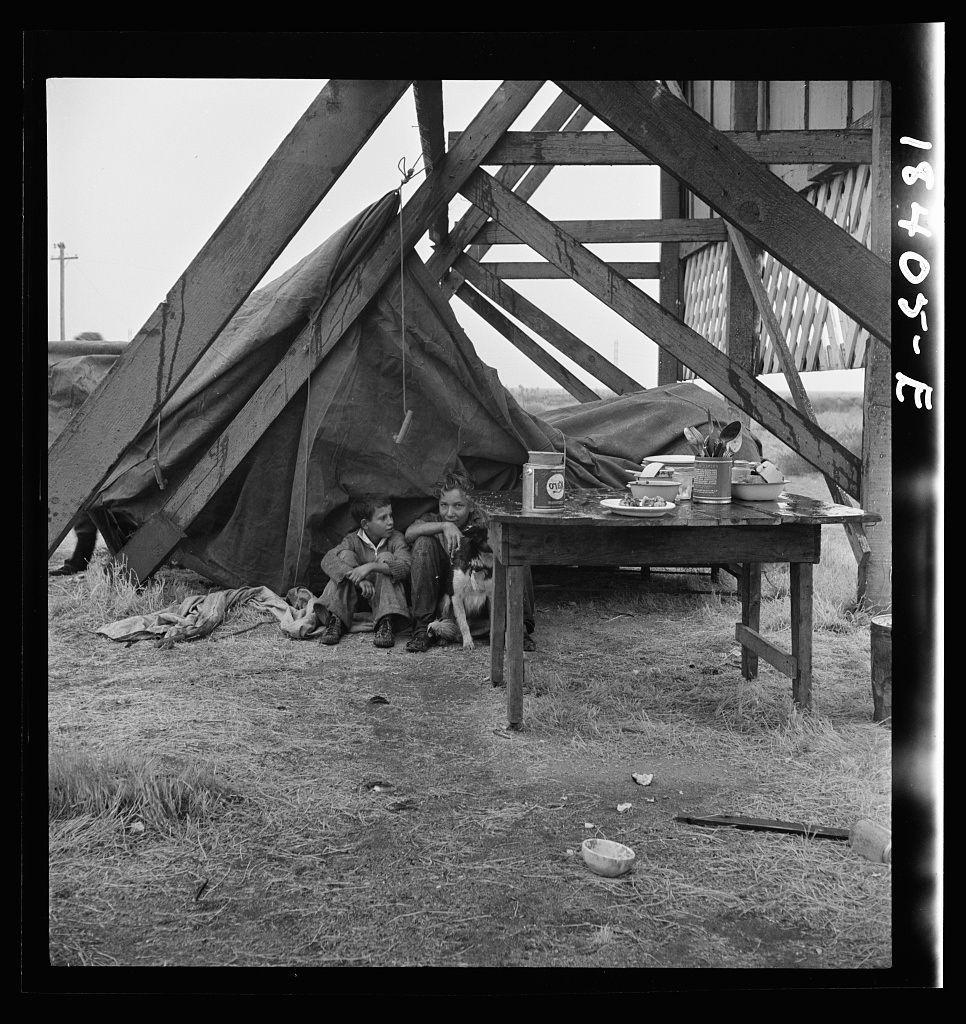 The image is a black and white photograph depicting what appears to be a group of people, possibly families or individuals living in an impoverished condition. They are situated outdoors under the shelter of makeshift structures that resemble tents constructed from tarps on wooden frames. The setting seems rudimentary with sparse belongings scattered around—a table laden with items like food containers, cups, and utensils suggests domestic activity despite the lack of proper facilities.

Several people can be seen; some sitting close together while others are lying down or seated farther apart. Their expressions convey fatigue or despair. There's a visible contrast between their modest living conditions and everyday objects that signify normalcy in daily life—tables with food, personal belongings. The ground is bare earth with patches of dried grass.

The photograph captures the stark reality of hardship without any overt action, but it communicates through stillness—the quiet resilience or acceptance possibly exhibited by those within this setting against a backdrop suggesting economic hardship or displacement.