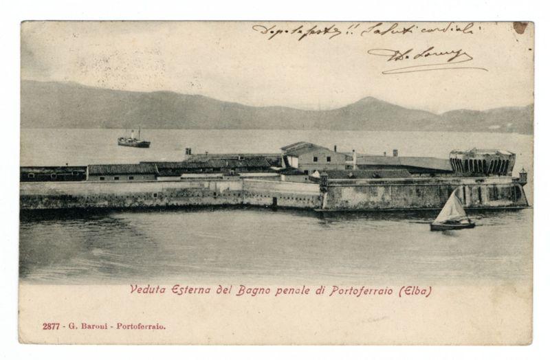 The image depicts an aged postcard showing a waterfront scene with prominent buildings and maritime activity. At the forefront, there's a large body of water reflecting light, suggesting either sunrise or sunset due to its soft glow on the surface. A sailboat is seen at rest in the lower right corner, indicating calm sea conditions.

Central to the composition are two elongated structures that appear industrial; one with several tiers and others having flat roofs. These buildings seem part of a harbor infrastructure, possibly for docking boats or storage purposes. The architecture suggests an older era, likely mid-19th century due to its style.

In the background, calm water extends towards hills in the distance under a clear sky with no visible clouds, giving the scene tranquility and space. There's another smaller vessel farther out on the left side of the postcard, reinforcing the maritime theme. The color palette is monochromatic, consistent with aged photographic prints or original imagery from around that time.

Text at the bottom provides context: "Veduta Esterna del Bagno penale di Portoferraio (Elba)" translates to "Exterior View of the Prison Penitentiary in Portoferro (Elba)." Additional information cites G. Baroumi, likely indicating the photographer or artist's name and suggests a connection with Elba island off Italy.

The top right corner includes signature [...]