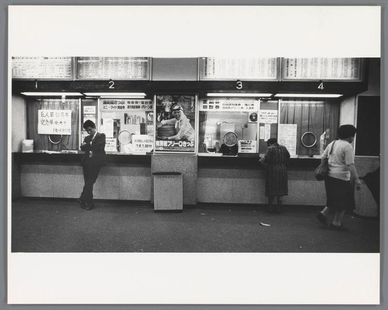 The image is a black and white photograph depicting an urban scene, possibly from the late 1960s or early 70s based on fashion styles. It shows four numbered service counters, each with various posters and signs in Japanese indicating services offered such as lottery tickets ("lottery," "lotto"), financial transactions (possibly related to savings), and other utilities like water bills for residents of Hibiya Ward. Individuals are seen interacting at the counters; one person is leaning on a counter reading a paper or book, another appears to be engaging with an attendant through a transparent window, while two others stand in line waiting their turn.

The environment suggests a public service office within Japan during a time when English was not commonly used for signage. The photograph captures candid moments of daily life and the social interaction typical at such services locations, reflecting on how people engaged with state-provided utilities or leisure activities like lottery tickets through these counters.