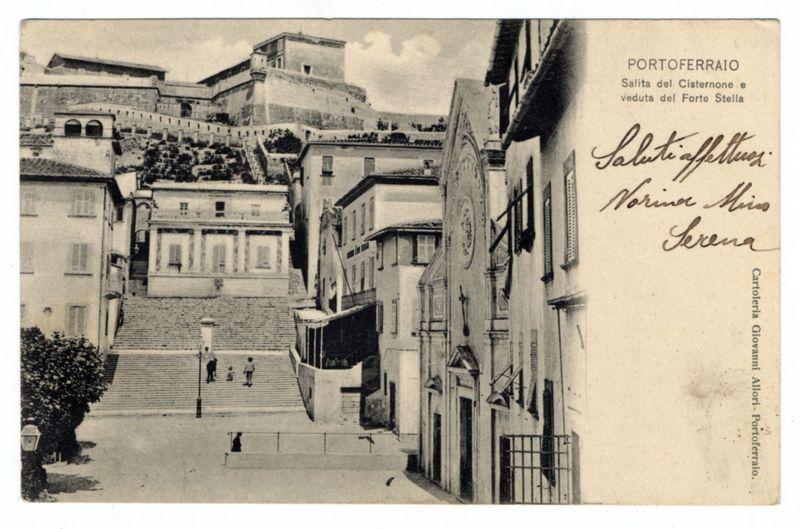 The image displays a monochromatic vintage postcard featuring architectural structures. The backdrop showcases an elevated, fortified building perched on what appears to be the crest of a hill or rocky outcrop, suggesting historical significance and possibly military importance as it overlooks its surroundings from such height.

Below this prominent fortress-like structure is a complex with several buildings that have classical European architecture elements like arched windows and decorative facades. Notably, there's an ornate clock tower on the right side of the image which seems to be part of another building or establishment adjacent to it.

In the foreground, we observe stairs leading up towards what could potentially be a town square or plaza with steps visible descending into view from above. A few individuals are seen at different levels: some walking down the stairs and others ascending them, providing scale and life to this historical setting. The presence of trees on both sides adds greenery contrasting against the built environment.

On top of the postcard is handwritten text that appears to be a greeting or signature followed by cursive writing which might indicate the sender's name or a location in Italian ('Salita del Cisternone e Veduta del Forte Stella'), hinting at its geographical origin. Additionally, there’s printed text along the right side identifyi [...]