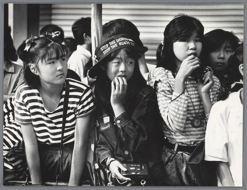 The image is a black and white photograph depicting four young Asian girls sitting closely together, seemingly waiting in line or at an event. The setting appears to be outdoors with buildings visible in the background behind them.

Starting from the left:
- One girl has her hair adorned with two bows on either side of her head.
- Next to her is a girl wearing striped clothing and carrying what seems like a bag strap over her shoulder, looking off into the distance with a neutral expression. She appears focused or contemplative.
- The third girl wears glasses, a dark jacket, denim overalls, and holds a small object in her hand that resembles a camera or perhaps food packaging. Her head is tilted slightly downwards as she looks at what's in her hands.

The fourth person on the right edge of the photo has their face partially obscured but appears to be wearing a patterned garment with sleeves rolled up to reveal stripes underneath, and they are looking away from the group towards something outside the frame.
All individuals exhibit casual postures typical of waiting or being engaged in quiet observation. The photograph captures a candid moment that conveys an atmosphere of anticipation or curiosity among these young people.

Additional information about this image can be found at: Groep meisjes in Japan, Ed van der Elsken, in or after c. 1980 - 1990
https://images.loener.nl [...]