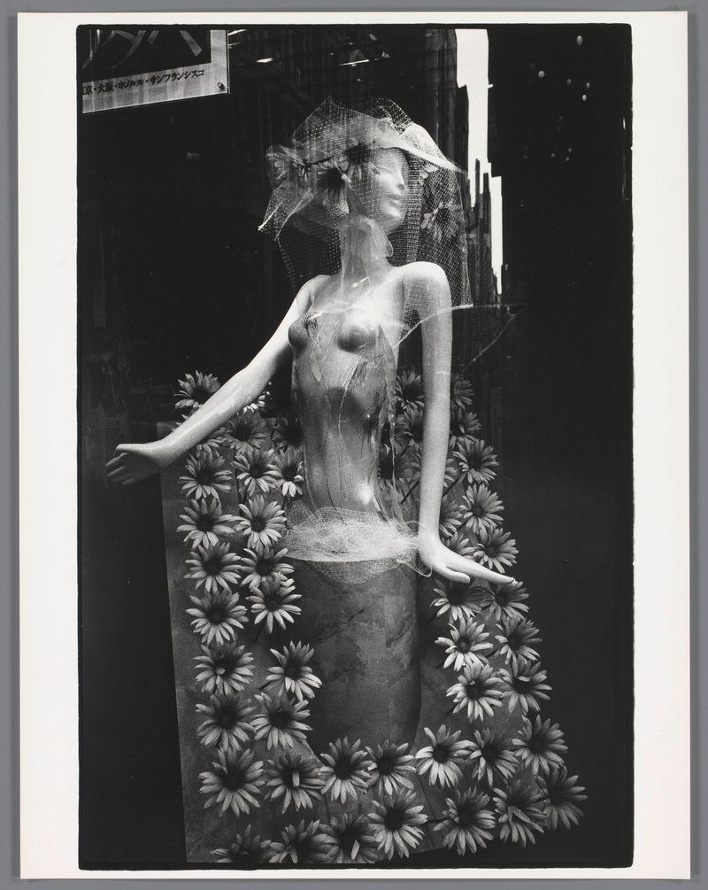 This image is a black and white photograph featuring a mannequin dressed in an elegant, sleeveless outfit with floral patterns. The mannequin has its arms extended outward, as if embracing the flowers that surround it, which appear to be daisies or sunflowers due to their shape and size. Overhead, there's what seems like netting material draped over the top of the head area, adding a whimsical touch to the composition.

The background is dark and nondescript, drawing focus towards the subject in the foreground. In one corner of the image, Japanese text can be seen on a signboard or banner, suggesting that this photograph was taken in Japan during Ed van der Elsken's visit there in 1959-1990.

The overall mood conveyed by this photo is nostalgic and artistic, capturing an evocative moment where fashion meets nature through the mannequin’s floral attire. The play of light and shadow on the figure enhances its ethereal quality.