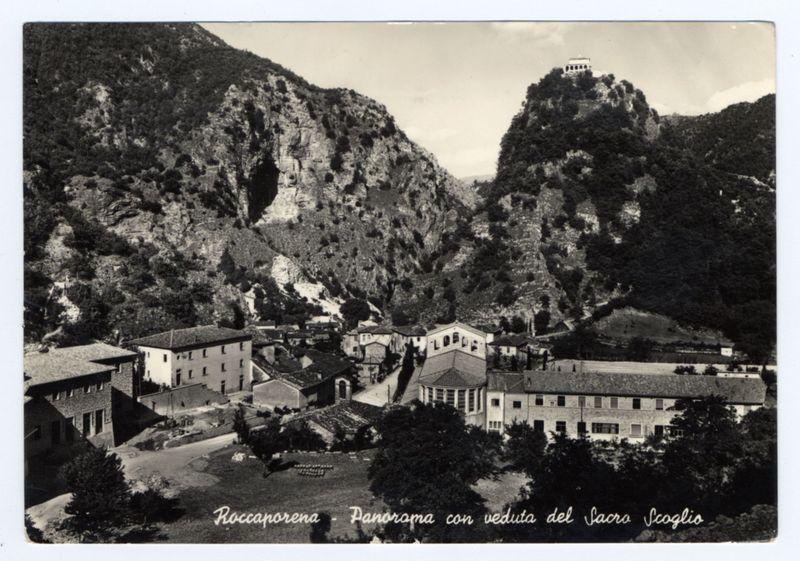 The image depicts a black-and-white vintage postcard featuring an aerial view of Roccaporena, specifically showcasing the town with its surrounding mountainous terrain and a prominent structure atop one peak. The mountains are rugged with visible rock faces and vegetation sparsely distributed across them. In the foreground is a small settlement comprising several buildings with traditional architecture; flat roofs on some houses and pitched roofs on others. One notable building has a distinctive bell tower, indicating it may be a church or religious site.

A paved road winds through the town, leading towards another large structure in the distance which appears to have multiple levels and balconies, possibly an observatory or historical edifice overlooking the area. The postcard bears text at the bottom: "Roccaporena - Panorama con veduta del Sacro Scoglio," suggesting it is a view of Roccaporena including that particular landmark.

The image captures the essence of early 20th-century photography, with its sepia tones and somewhat grainy texture. It gives an impression of historical significance or tourism appeal for this Italian town nestled in picturesque mountain scenery.