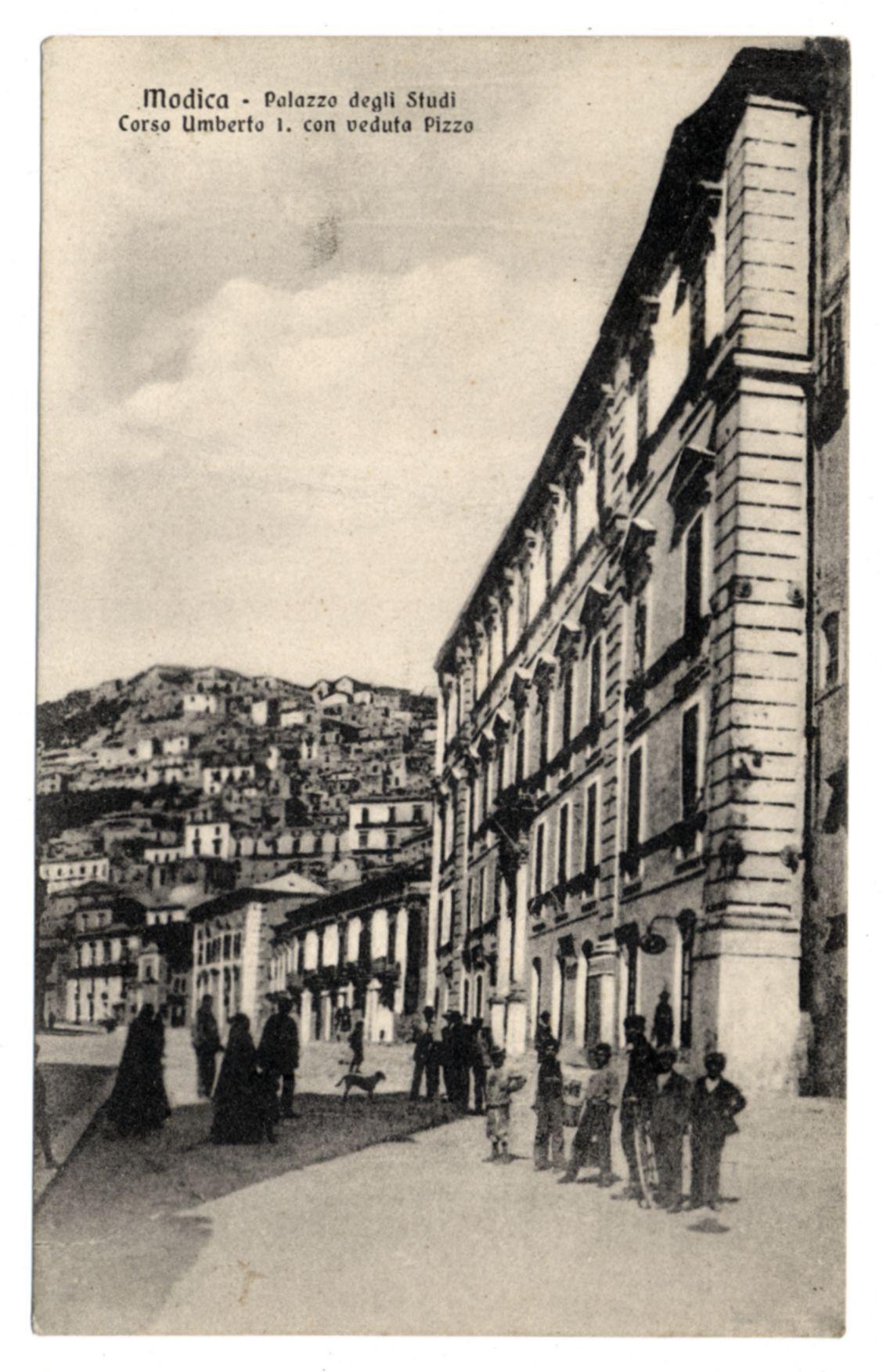 The image is a black and white historical postcard depicting the Palazzo degli Studi on Corso Umberto I in Modica, with views of Piazza. The photograph captures an urban street scene during what appears to be early 20th-century times. A row of classical architecture lines one side of the street while pedestrians are seen strolling along or standing and conversing near a fountain located at the entranceway of the Palazzo.

In the foreground, several individuals dressed in period attire can be observed: women wearing long dresses with dark skirts and light-colored blouses, men sporting suits and hats indicative of the era. There is also a dog accompanying one individual. The architecture features multiple levels, arched windows, and balconies characteristic of early 20th-century European urban design.

The background reveals hilly terrain dotted with houses or buildings suggesting a hillside town setting. Above it all, an overcast sky hangs above the scene. A caption on top of the postcard reads "Modica - Palazzo degli Studi Corso Umberto I., con veduta Piazza," indicating that this image is likely from Modica and shows both the Palace itself as well as a view across to another street called 'Piazza.'
