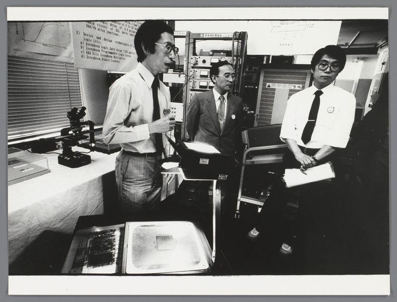 The image depicts three men in a laboratory setting. The man on the left is standing and holding what appears to be a small electronic device or component, examining it closely with focused attention. He wears glasses, has dark hair styled away from his face, and sports a tie over an open-collar shirt.

In the center stands another gentleman who seems engaged in conversation; he gestures toward something off-camera while speaking. This individual is wearing a suit with a badge pinned to his lapel and sporting spectacles as well.

The man on the right appears more relaxed than his companions but still attentive, holding what could be some papers or documents. He has short hair and wears glasses similar in style to those of the other two men.

Behind them are various pieces of electronic equipment including a microscope mounted above a tray with samples, racks filled with different devices possibly for testing purposes, and an array of wires leading into metal boxes that suggest circuitry work is taking place here. The overall atmosphere suggests an educational or professional setting, likely part of the excursion mentioned in the caption.

In terms of context provided by the URL linked to this image, it appears these individuals are involved with a group from Groningen visiting Japan as part of an educational trip and they're currently at a laboratory where technical demon [...]