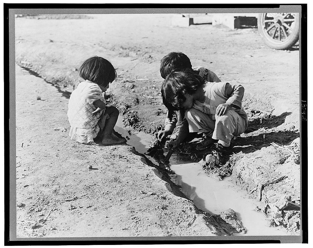 The image is a black and white photograph depicting three young children engrossed in play near an open ditch. The setting appears rural, with sandy soil suggesting arid conditions or agricultural surroundings. In the background, there's evidence of human habitation like a wooden structure and a large vehicle wheel, hinting at nearby infrastructure.

The child on the left is crouched down closely inspecting something within the ditch, possibly rocks or mud, while extending their arm into it as if searching for hidden objects. The middle child mirrors this posture but seems to be more focused on reaching inside with both hands and appears slightly bent over in concentration. The third child on the right has one knee touching a muddy bank of the ditch, observing something intently within the waterlogged area.

The clothing worn by all children is indicative of a time period where such attire might have been common; they appear to be wearing loose-fitting garments suitable for play and outdoor activities in warm climates. The overall atmosphere conveys innocence and curiosity as these young individuals explore their environment through casual, unstructured means without any modern toys or electronic devices visible.

There's an air of simplicity and focus on the natural elements around them, reflecting a moment captured during childhood exploration with little to no indicati [...]